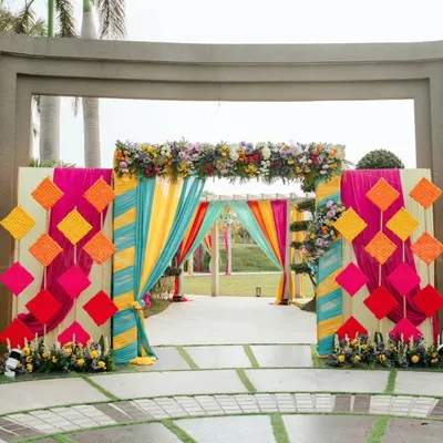 A grand entrance that screams celebration. The mix of colorful drapes in yellow, teal, and pink with geometric hangings and floral arrangements sets a festive tone from the moment guests arrive for the mehendi.