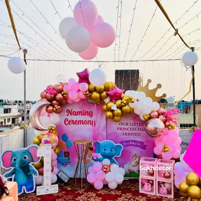 A grand and festive rooftop setup for a princess-themed naming ceremony. The decor is built around a storybook-style backdrop and features a balloon arch in pink, white, and gold, character cutouts, and a light-up number '1'.