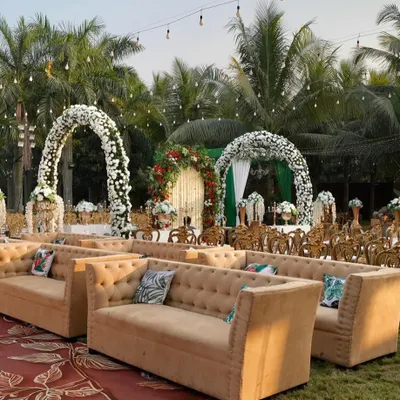 An outdoor daytime reception setup with elegant lounge seating. The white floral arches and lush palm trees create a beautiful, relaxed atmosphere for guests to mingle and celebrate.