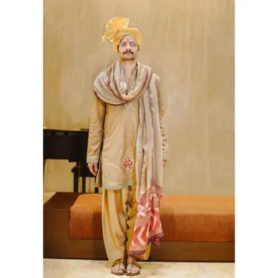 A classic, full-length portrait of the groom. His confident stance and traditional attire, complete with a vibrant yellow turban, are captured against a simple, elegant backdrop.