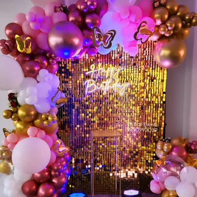 A vibrant birthday backdrop featuring a gold sequin shimmer wall. The wall is framed by an elaborate balloon garland in shades of pink, white, and gold, and is finished with a 'Happy Birthday' neon sign.