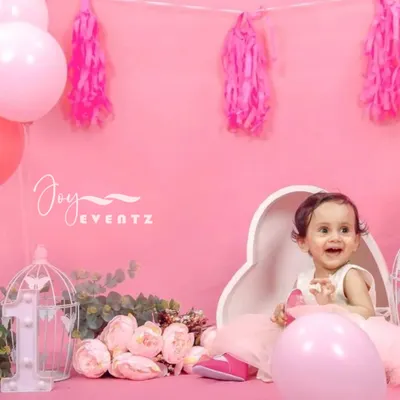 This adorable cake smash photoshoot for a baby girl features a solid pink backdrop with matching balloons and floral props. The heart-shaped seat and decorative birdcage add a charming touch to capture this special milestone.