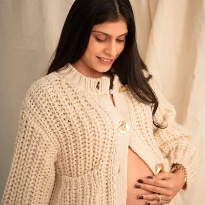 A serene solo portrait from a cozy studio session. The mom-to-be, wearing a chunky knit cardigan, cradles her bare belly with a gentle smile.