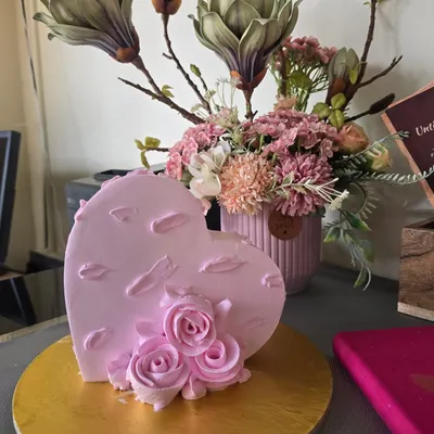 Hand-sculpted roses and petal-like textures finish our blush pink heart cake, displayed upright beside flowers at the counter.