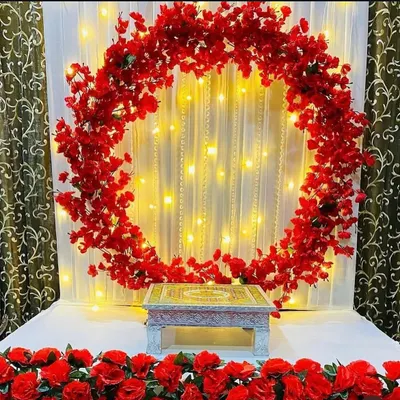 A beautiful circular frame of red flowers set against a white curtain with pearl strings and fairy lights. This setup is ready for your idol, providing a vibrant and glowing backdrop.