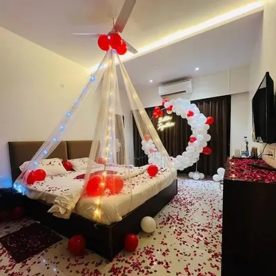 A complete room transformation featuring a circular balloon arch with a neon sign, a fairy light canopy, and a floor covered in rose petals. This red and white theme is a popular choice for romantic anniversary and birthday celebrations at home.