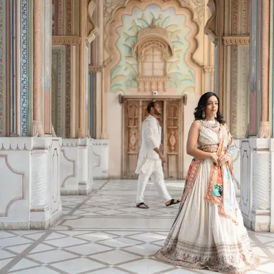 A portrait of modern royalty in a Jaipur palace. The bride stands poised in an elegant lehenga, while her partner walks in the background, creating a sense of depth and story within the frame. This is a perfect example of a destination heritage shoot.