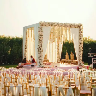 An outdoor wedding mandap with a clean, modern design. The structure is adorned with white flowers and flowing drapes, creating an elegant and timeless look for a daytime ceremony.