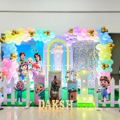 A bright and colorful Cocomelon birthday party setup. This decoration includes multiple character cutouts, a pastel rainbow balloon arch, and a large light-up number '1'.