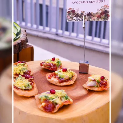 Our Avocado Sev Puri is a modern take on a classic chaat. Crispy puris are topped with fresh avocado, tangy chutneys, and pomegranate seeds, served on a rustic wooden platter.