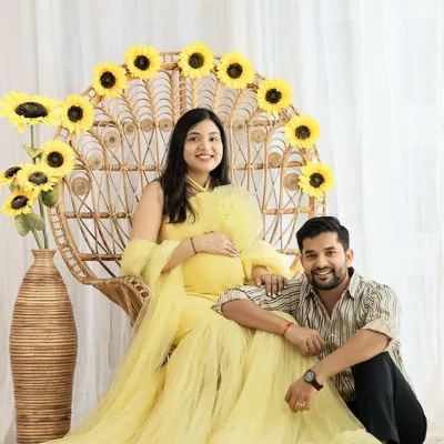 A bright and joyful couple portrait. The mom-to-be is radiant in a yellow tulle gown, seated on a peacock chair decorated with sunflowers, while her partner sits lovingly by her side.