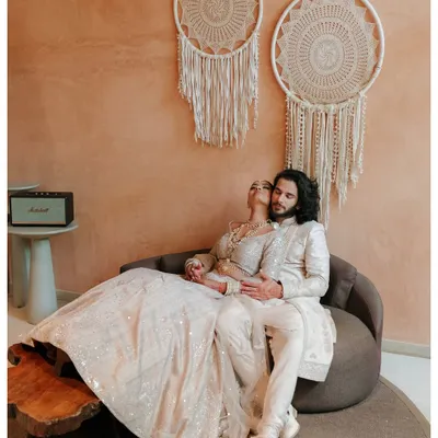 A stunning indoor portrait with the couple dressed in elegant white traditional wear. The bohemian decor and warm lighting create a sophisticated and romantic atmosphere.