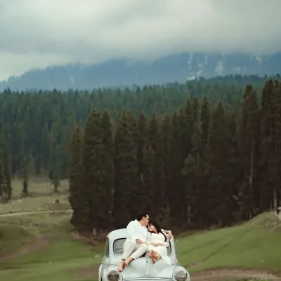 A breathtaking shot of a couple sharing an intimate moment on a vintage car, surrounded by the vast, pine-covered hills of Kashmir. This is the essence of a destination pre-wedding shoot, combining romance with awe-inspiring scenery.
