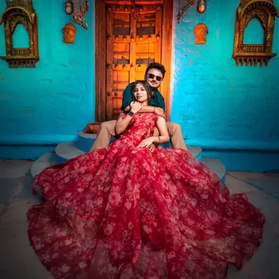 A vibrant and artistic pre-wedding shot. The red floral printed organza gown looks stunning against the colorful blue wall, creating a photo that is full of life and personality.