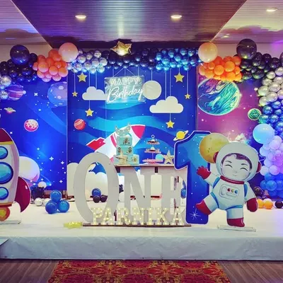An out-of-this-world space theme for a first birthday. The stage is filled with a galaxy backdrop, a rocket ship prop, an astronaut cutout, and balloon garlands representing planets and stars.