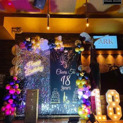 A glamorous "Cheers to 18 Years" birthday party setup. This decor features a silver sequin wall, a custom backdrop, a 'Happy Birthday' neon sign, and light-up marquee numbers, perfect for a milestone celebration.