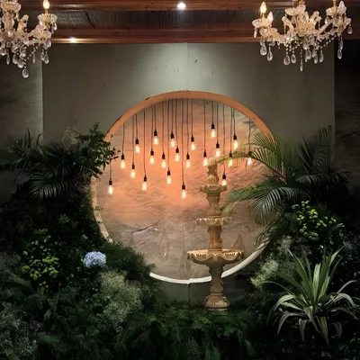 A romantic photo backdrop from our tropical-themed engagement. A classic fountain is framed by lush jungle foliage and a circular installation of warm Edison bulbs, creating a magical, moody setting under elegant crystal chandeliers.