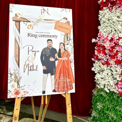 The final printed welcome board for Pooja and Nikhil's ring ceremony, set up on an easel at the event venue. It looks stunning next to the floral decorations.