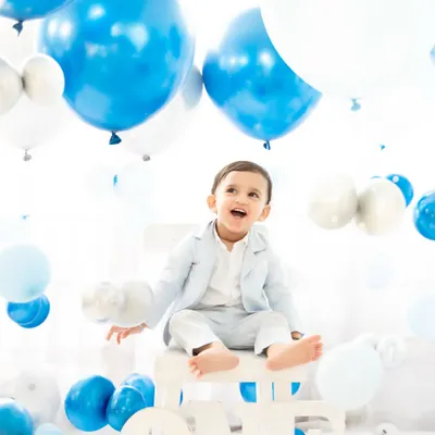 A baby boy celebrates his first birthday in a classic blue and white balloon-filled setup. His joyful expression and the festive atmosphere perfectly capture the happiness of this major milestone.
