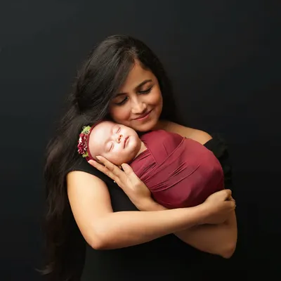 A mother with long dark hair, dressed in black, holds her sleeping newborn close to her chest. The baby is swaddled in a deep red wrap, creating a beautiful color contrast against the dark backdrop.
