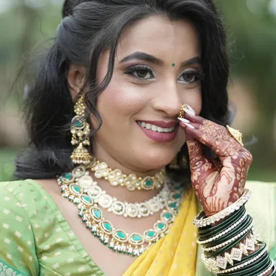 A happy bride adjusts her nath, giving a beautiful view of her layered Kundan necklace set with sea-green stones and matching jhumka earrings.