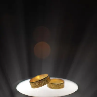 A creative shot of the wedding rings, spotlit against a dark background. The dramatic lighting makes the gold bands shine, symbolizing their precious commitment.