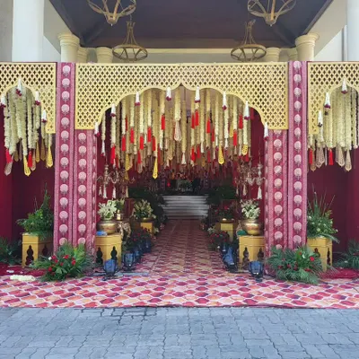A grand and traditional welcome gate for a palace wedding. The entrance is designed with a Rajasthani-style arch, red drapes, and hanging floral tassels made of marigolds and tuberoses.