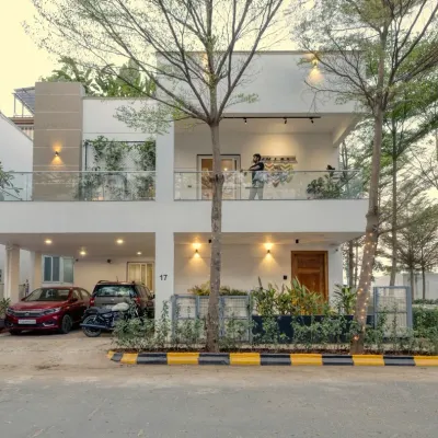 The facade of Villa 17 in Bangalore. This two-story home features a clean white exterior, a glass balcony railing, and strategically placed lighting, softened by a ledge of green creepers for a touch of nature.