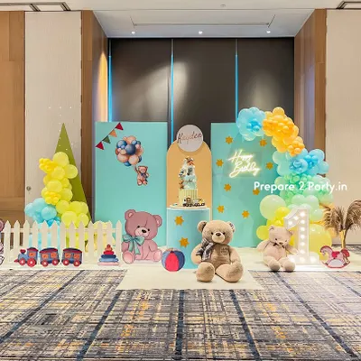 A cheerful and playful first birthday party setup in a large hall. The theme features pastel blue and yellow backdrops, a white picket fence, cute teddy bear cutouts, and a light-up marquee number "1".