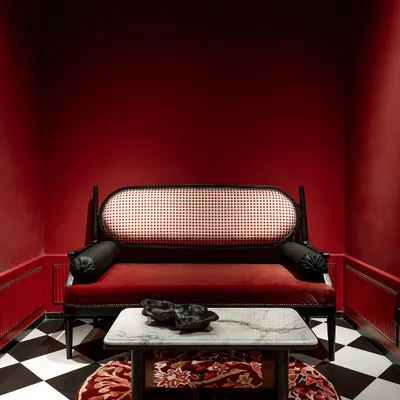 A statement piece from our House of Goya collection. This classic daybed features a black-lacquered frame, a cane back, and rich red velvet upholstery. It's set in a dramatic, color-drenched room with a checkerboard floor.