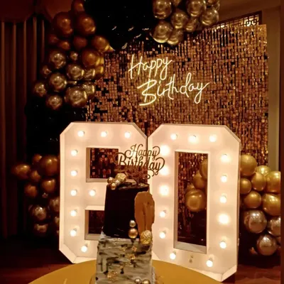 This 60th birthday photo corner was designed for maximum impact. It features a gold sequin shimmer wall, a 'Happy Birthday' neon sign, and oversized light-up '60' numbers, complemented by a classic black and gold balloon garland.