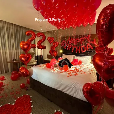 A classic romantic birthday room decoration in a hotel suite. The setup features red heart-shaped foil balloons, number balloons indicating the age, "Happy Birthday" lettering above the bed, and a floor decorated with rose petal hearts.
