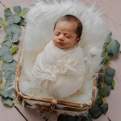A perfect little bundle of joy. This newborn is peacefully asleep in a rustic basket, swaddled in white on a fluffy fur blanket and surrounded by eucalyptus leaves.