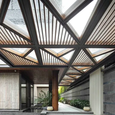 The entrance canopy of the Bammi Residence, with its intricate geometric design, filters sunlight to create a dynamic play of light and shadow. This architectural statement sets the stage for a home where every detail is considered, blending structural form with natural elements to craft a unique sense of arrival.