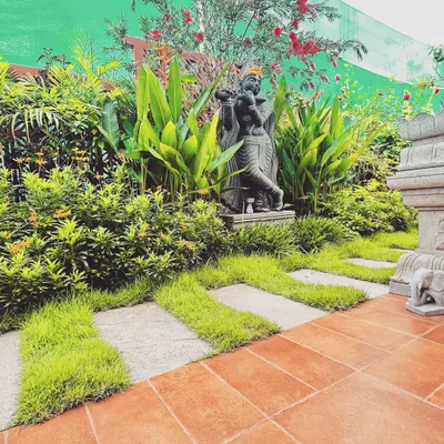 A statue of Krishna playing the flute is nestled among lush tropical foliage and vibrant red flowers. The pathway, made of stone pavers interspersed with grass, leads to this spiritual alcove, creating a sacred pause within the garden.