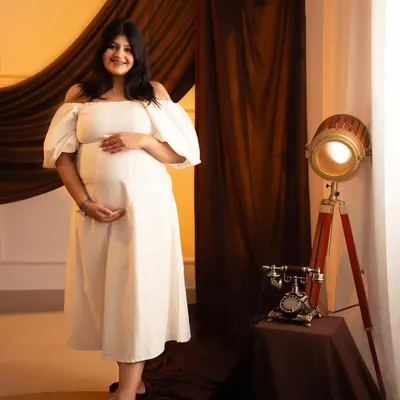 A classic studio portrait with a vintage touch. The mom-to-be looks radiant in a white off-the-shoulder dress, complemented by the warm lighting and antique props.