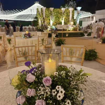 A close-up of a classic and elegant centerpiece. A pillar candle in a glass hurricane lamp is surrounded by a delicate arrangement of lavender roses, baby's breath, and daisies, all set on a mirrored base.