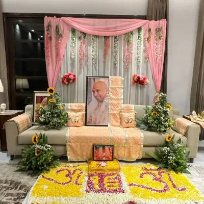 This is a complete view of a Guruji Satsang I organized. The main seating area features a portrait of Guruji, decorated with soft pink drapes and floral arrangements. In front, a large, intricate floral rangoli with the 'Om' symbol creates a powerful focal point for meditation and prayer.