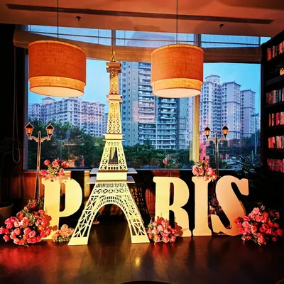 A stunning Paris-themed setup against a city skyline. The decor features a large Eiffel Tower cutout, 'PARIS' letters, and beautiful floral arrangements, perfect for a chic and sophisticated event.