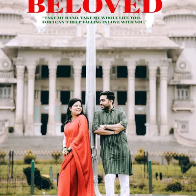 A grand, symmetrical shot of a couple in traditional wear standing before the Vidhana Soudha in Bangalore, framed as a 'Beloved' movie poster.