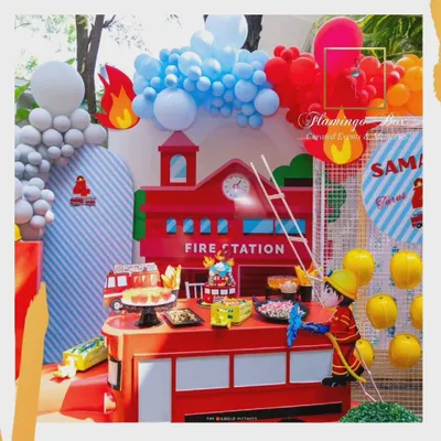 The main setup for a firefighter-themed birthday, featuring a custom fire station backdrop and a dessert table designed to look like a fire truck. Balloon arrangements in red, blue, and orange mimic fire and water.