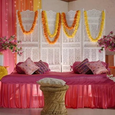 A classic Mehendi seating arrangement featuring a comfortable divan in bright pink, set against a traditional white carved screen and decorated with marigold garlands.