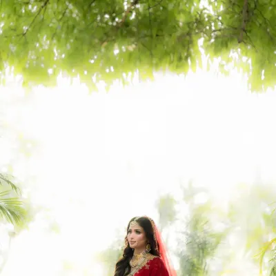 A bride framed by nature, her red lehenga glowing against the soft, diffused light. This is a portrait of pure, ethereal beauty.