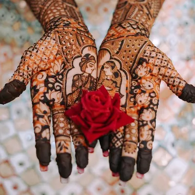 A close-up of the bride's hands showcasing the incredible detail of her mehndi, featuring portraits of the couple. A small red rose adds a beautiful pop of color.