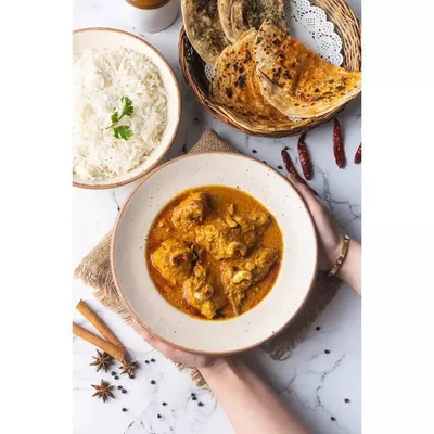 A light and airy composition of Chicken Korma, served with rice and roti. The hands holding the bowl add a human element, making the meal feel warm and inviting.