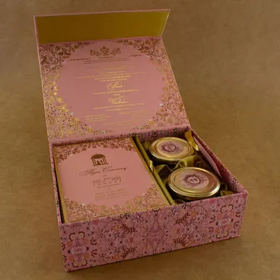 An exquisite pink and gold boxed invitation, perfect for a grand wedding or engagement. The box opens to reveal the invitation details printed on the inside of the lid, with compartments for the card inserts and two jars for sweets or dry fruits.