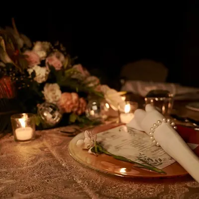 A beautifully set table for an intimate dinner. The place setting features a personalized menu, a pearl napkin ring, and a single flower, all glowing in the warm light of a nearby candle.