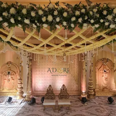 A magnificent golden-themed mandap for a traditional Muhurtham ceremony. The design features a lattice of white flowers on the ceiling, ornate pillars, and idols of deities, creating a sacred and regal space.