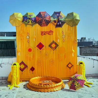 This is a classic Haldi ceremony setup I did on a sunny rooftop. The backdrop features bright yellow drapes with the word 'Haldi' in Hindi, decorated with colorful Rajasthani umbrellas and traditional hangings for a festive look.