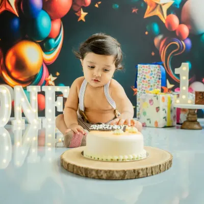 The moment of anticipation just before the smash. This little one is eyeing his first birthday cake in a festive setup with 'ONE' marquee lights and colorful balloons.